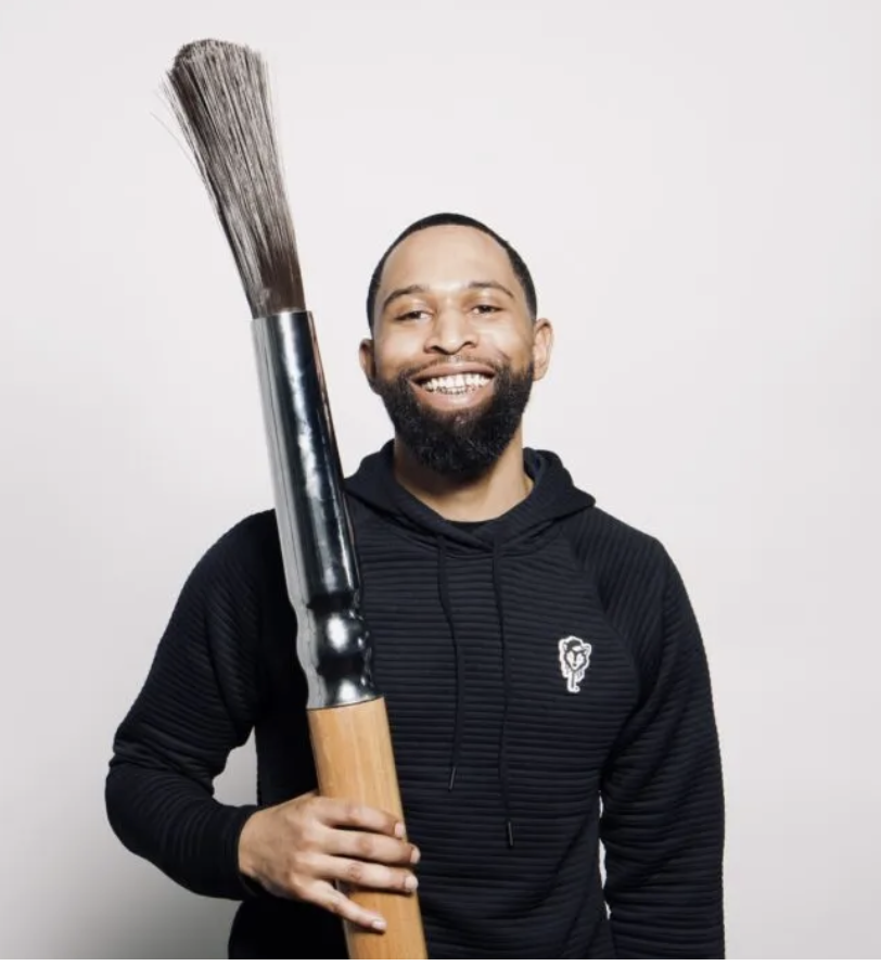 Smiling man with a beard wearing a black hoodie, holding an oversized paintbrush against a plain background.